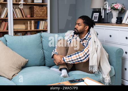 Junger deprimierter Afroamerikaner, der mit gekreuzten Armen auf dem Sofa sitzt. Rassismus-Opfer für Hautfarbe schwarz männlich mit Depressionen nach dem sein Stockfoto