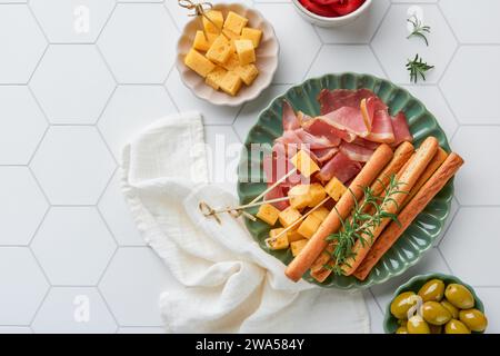 Schinkenscheiben oder Marmeladen. Antipasti. Köstliche grissini-Sticks mit Schinken, Käse, Rosmarin, Oliven auf grünem Teller auf weißem Betonhintergrund Stockfoto