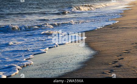 Ein Sandstrand mit Wellen, die an die Küste kommen und das Licht der untergehenden Sonne reflektiert vom Wasser. Stockfoto