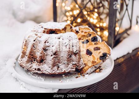 Traditioneller Weihnachtskuchen oder Obstkuchen mit getrockneten Früchten auf Teller mit ausgeschnittenen Stücken, Schnee und Weihnachtsbeleuchtung auf Hintergrund. Stockfoto