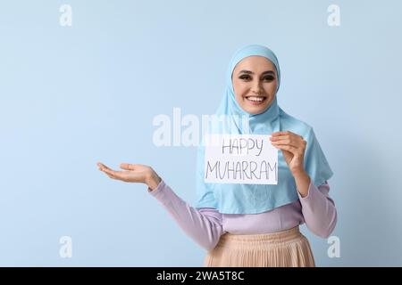 Junge muslimische Frau, die Papier mit Text HAPPY MUHARRAM auf blauem Hintergrund hält Stockfoto