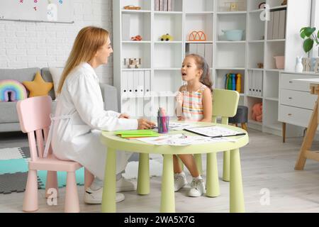 Niedliches kleines Mädchen mit logopädischer Sonde und Logopädie, die einen Brief am Tisch im Büro ausspricht Stockfoto