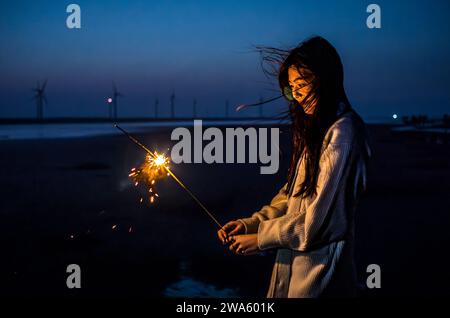 Taichung, China. Januar 2024. Eine junge Frau hält am 01/2024 in Taichung, Taiwan, einen Funkeln an den Ufern der Gaomei Wetlands. Die Feuchtgebiete, ein 300 Hektar großes Naturschutzgebiet, das für seine Artenvielfalt und landschaftliche Schönheit bekannt ist, bieten eine ruhige Kulisse, während der Tag mit Windturbinen endet, die von Wiktor Dabkowski vor dem pulsierenden Himmel geschildert werden. dpa/Alamy Live News Stockfoto