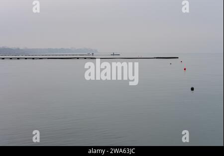 Grado Meer an einem Wintertag mit einem dicken Nebel, der die Umgebung suggestiv und mystisch macht. Foto vom Strand mit Leuten, die auf dem Pier laufen. Stockfoto