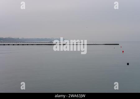 Grado Meer an einem Wintertag mit einem dicken Nebel, der die Umgebung suggestiv und mystisch macht. Foto vom Strand mit Leuten, die auf dem Pier laufen. Stockfoto