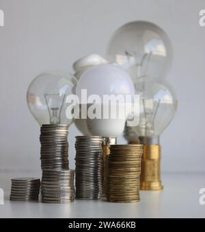 LED-Lampe und Glühlampen hinter den Münzstapeln isoliert auf weiß. Konzeptfoto zur metaphorischen Erhöhung der Energieeinsparung Stockfoto