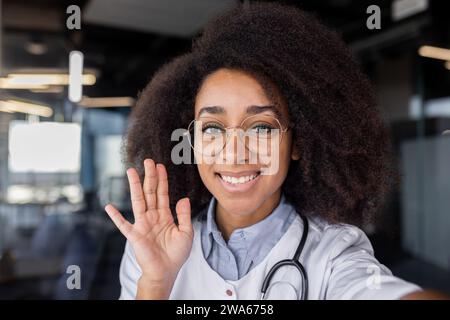 Nahporträt einer jungen, lächelnden afroamerikanischen Ärztin, die im Klinikbüro steht, mit der Hand vor der Kamera spricht, berät und grüßt. Stockfoto