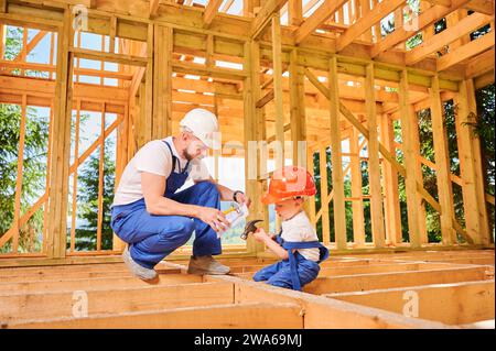 Vater mit Kleinkind Sohn baut ein Holzrahmenhaus. Ein Baumann, der seinem Sohn die Technik beibringt, auf der Baustelle Nägel mit Hammer zu schlagen, mit Helmen und blauen Overalls. Stockfoto