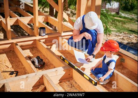 Vater mit Kleinkind-Sohn, der ein Haus mit Holzrahmen baut. Männlicher Baumeister zeigt seinem Sohn den Bauplan, trägt Helme und blaue Overalls an sonnigen Tagen. Zimmerei und Familienkonzept. Stockfoto