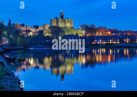 Blick in der Abenddämmerung auf Warkworth Castle und Warkworth Village, die sich im Fluss Coquet spiegeln, wie man von einem Spaziergang mit Warkworth Castle im Flutlicht sehen kann. Stockfoto