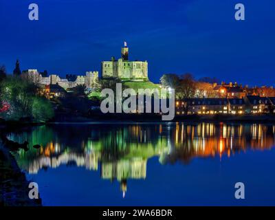 Blick in der Abenddämmerung auf Warkworth Castle und Warkworth Village, die sich im Fluss Coquet spiegeln, wie man von einem Spaziergang mit Warkworth Castle im Flutlicht sehen kann. Stockfoto