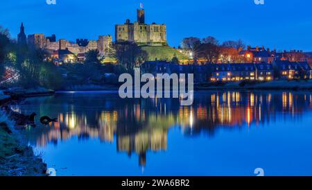 Blick in der Abenddämmerung auf Warkworth Castle und Warkworth Village, die sich im Fluss Coquet spiegeln, wie man von einem Spaziergang mit Warkworth Castle im Flutlicht sehen kann. Stockfoto