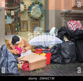 Unten und draußen in Rom sitzt eine Obdachlose mit ihren Besitztüchern in Plastiktüten vor einem Geschäft mit „Frohe Weihnachten“ auf einer Straße in der Vatikanstadt. Stockfoto