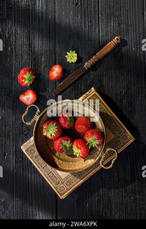 Frische Erdbeeren in Vintage Bronzeschale auf einem alten alten Buch auf dunklem hölzernem Hintergrund mit seitlicher Beleuchtung Stockfoto