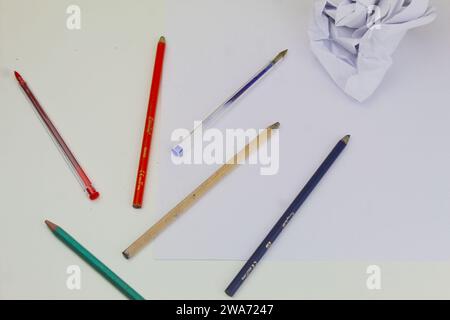 Ein Foto von Malstiften eines Schülers und weißen Papierstücken, die auf einem weißen Schreibtisch zusammengerollt sind. Stockfoto
