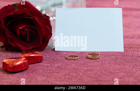 valentinstag, Verlobungsringe, rote Rose, zwei herzförmige Pralinen und eine weiße Karte mit Kopierraum Stockfoto