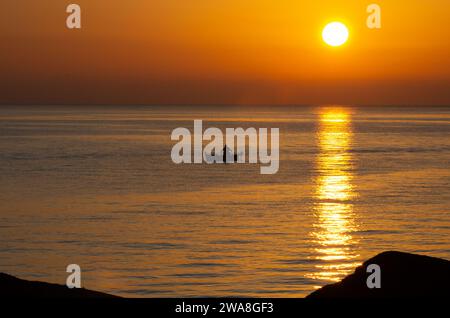 Der Sonnenaufgang flüstert, während ein einsamer Fischer, eine auffällige Silhouette, sein Boot navigiert und in den Morgentönen auf der ruhigen Leinwand des plätschernden Meeres tanzt. Stockfoto