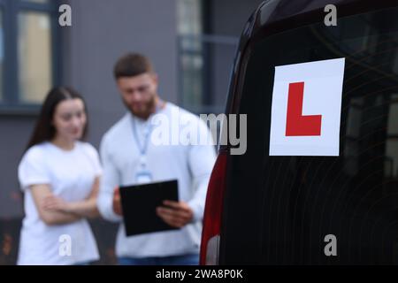 Fahrer und Kursleiter mit Klemmbrett in der Nähe des Autos im Freien, selektiver Fokus auf L-Platte. Fahrschule Stockfoto