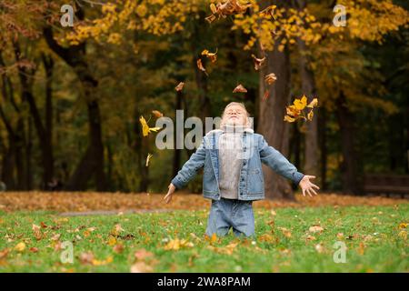 Glücklicher Junge, der mit trockenen Blättern auf grünem Gras im Herbstpark spielt Stockfoto