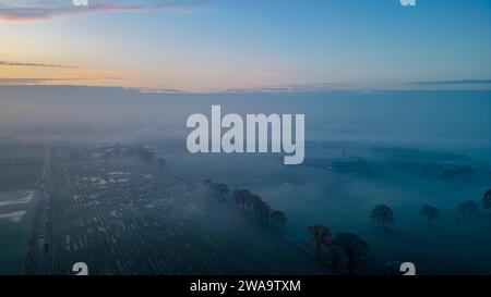 Dieses Bild fängt den atemberaubenden Moment des Sonnenaufgangs über einem riesigen Ackerland ein, das von Morgennebel umgeben ist. Das sanfte Licht der aufgehenden Sonne färbt den Himmel mit zarten Rosa- und Blautönen, während der Boden darunter ein Wandteppich aus schattigen Feldern und Bäumen aus dem Nebel ist. Der Nebel erzeugt eine traumhafte Qualität und verschwimmt die Grenzen zwischen Land und Himmel. Die verstreuten Bäume stehen als Silhouetten vor der trüben Kulisse, und der ferne Horizont deutet auf das Versprechen eines neuen Tages hin. Dieser Blick aus der Luft bietet eine friedliche und außerirdische Perspektive auf die Dämmerung und lädt zur Betrachtung des Tagess ein Stockfoto