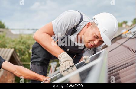 Männer montieren Photovoltaikmodule auf dem Dach des Hauses. Nahaufnahme Elektriker im Helm Blick auf Ebenheit des Solarpanels während der Installation. Konzept der alternativen und erneuerbaren Energien. Stockfoto