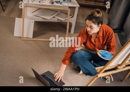 Künstlerin, die in ihrem Atelier sitzt, Laptop-Computer benutzt, Videogespräche mit Studenten, Online-Kunstunterricht, arbeitet an einem Projekt, Malerin forscht. Stockfoto