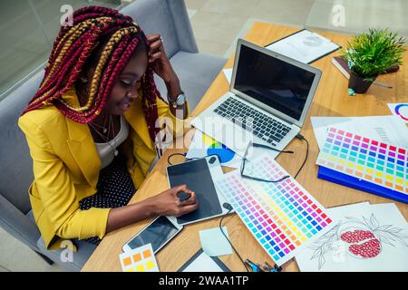 Eine Modedesignerin mit Afro-Pigtails Dreadlocks, die in ihrem Atelier oder Druckverlag arbeitet, und Grafikdesignerin zeichnet eine Skizze auf einem Tablet Stockfoto