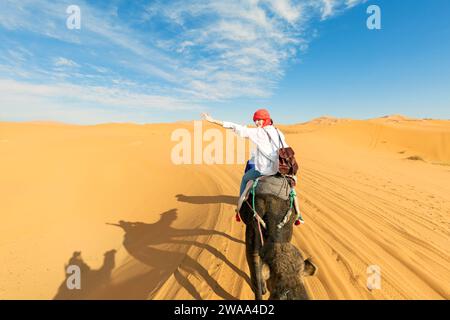 Eine junge Frau reitet auf einem Kamel durch die Dünen in der Sahara. Blick auf die Frau von hinten, im Hintergrund, kleine Silhouetten einer anderen Tour Stockfoto