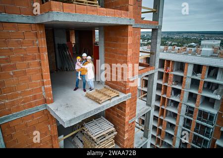 Frau umarmt Ehemann und Kind, während sie auf Betonboden in einem Wohngebäude steht, das gerade gebaut wird. Familie posiert in einem neu errichteten Appartementgebäude. Drohnenansicht Stockfoto