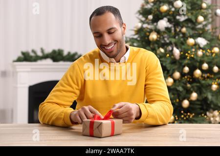 Glücklicher Mann, der das Weihnachtsgeschenk am Holztisch im Zimmer öffnet Stockfoto