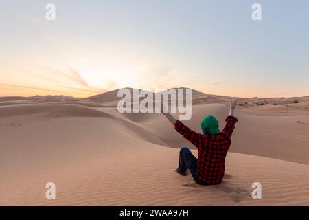 Ein junger Mann mit offenem Arm, der auf den Wüstendünen sitzt - Reisekonzept Stockfoto