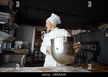 Glückliche junge Bäckerin, die eine Metallschale mit Sauerteig hält Stockfoto