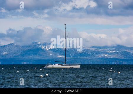 Ein Segelboot ankerte nach einem Schneesturm im späten Frühjahr am Lake Tahoe, mit bewölktem Himmel und frischem Neuschnee auf den Bergen im Hintergrund. Stockfoto