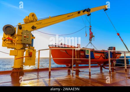 Orangefarbenes Rettungsboot auf großem Frachtschiff und Kran. Man-über-Bord-Übung. Rettungsboottraining. Stockfoto