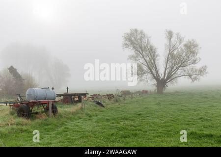 Ein einsamer Baum am Rande einer Wiese, der von Frühlingsnebel verschwimmt ist. Verschiedene landwirtschaftliche Maschinen am Zaun. Stockfoto