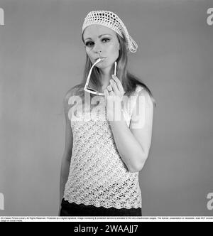 In den 1960er Jahren Eine junge Frau in einem Fotografen-Studio fotografierte mit einem gestrickten ärmellosen Pullover, der in einer luftigen, durchsichtigen Art gestrickt wurde. Die Kappe ist im selben Herrenhaus gestrickt. Sie trägt eine Sonnenbrille. 1967. Stockfoto