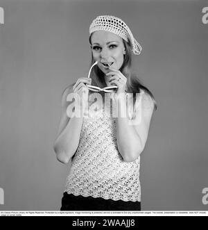 In den 1960er Jahren Eine junge Frau in einem Fotografen-Studio fotografierte mit einem gestrickten ärmellosen Pullover, der in einer luftigen, durchsichtigen Art gestrickt wurde. Die Kappe ist im selben Herrenhaus gestrickt. Sie trägt eine Sonnenbrille. 1967. Stockfoto