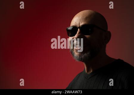 Hübscher, kahlköpfiger, bärtiger Mann mit Sonnenbrille, lächelnd und selbstbewusst. Isoliert auf rotem Hintergrund. Stockfoto