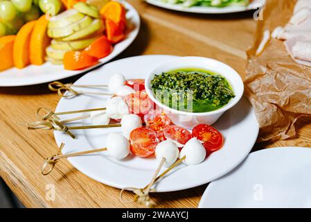 Frische Caprese Salatspieße mit Reifen Kirschtomaten, Mozzarella-Kugeln und einer Schüssel mit grüner Pesto-Sauce auf einem rustikalen Holztisch. Stockfoto