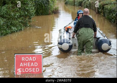 Worcester, 3. Januar 2024: Jack, siebeinhalb Jahre alt, wird mit dem Boot zu seinem Haus im englischen Dorf Severn Stoke gebracht, das durch Überschwemmungen vollständig abgeschnitten wurde, nachdem der Fluss Severn aufgrund anhaltender Regenfälle durch Sturm Henk seine Ufer geplatzt hatte. Der Rose and Crown Pub konnte gesehen werden, wie er Wasser herauspumpte, als er mit seinen selbstgemachten Hochwasserschutzanlagen um trocken kämpfte. - Credit: Stop Press Media/Alamy Live News Stockfoto