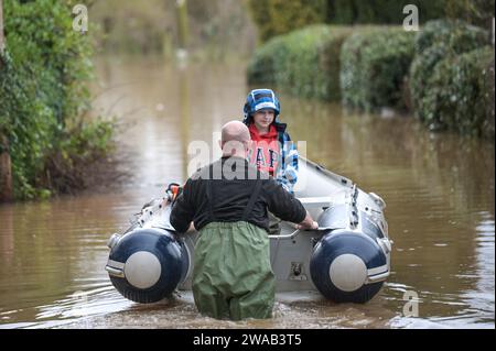 Worcester, 3. Januar 2024: Jack, siebeinhalb Jahre alt, wird mit dem Boot zu seinem Haus im englischen Dorf Severn Stoke gebracht, das durch Überschwemmungen vollständig abgeschnitten wurde, nachdem der Fluss Severn aufgrund anhaltender Regenfälle durch Sturm Henk seine Ufer geplatzt hatte. Der Rose and Crown Pub konnte gesehen werden, wie er Wasser herauspumpte, als er mit seinen selbstgemachten Hochwasserschutzanlagen um trocken kämpfte. - Credit: Stop Press Media/Alamy Live News Stockfoto