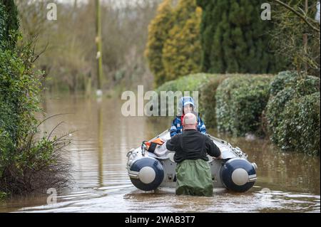 Worcester, 3. Januar 2024: Jack, siebeinhalb Jahre alt, wird mit dem Boot zu seinem Haus im englischen Dorf Severn Stoke gebracht, das durch Überschwemmungen vollständig abgeschnitten wurde, nachdem der Fluss Severn aufgrund anhaltender Regenfälle durch Sturm Henk seine Ufer geplatzt hatte. Der Rose and Crown Pub konnte gesehen werden, wie er Wasser herauspumpte, als er mit seinen selbstgemachten Hochwasserschutzanlagen um trocken kämpfte. - Credit: Stop Press Media/Alamy Live News Stockfoto