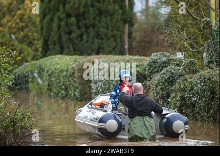 Worcester, 3. Januar 2024: Jack, siebeinhalb Jahre alt, wird mit dem Boot zu seinem Haus im englischen Dorf Severn Stoke gebracht, das durch Überschwemmungen vollständig abgeschnitten wurde, nachdem der Fluss Severn aufgrund anhaltender Regenfälle durch Sturm Henk seine Ufer geplatzt hatte. Der Rose and Crown Pub konnte gesehen werden, wie er Wasser herauspumpte, als er mit seinen selbstgemachten Hochwasserschutzanlagen um trocken kämpfte. - Credit: Stop Press Media/Alamy Live News Stockfoto