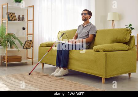 Blinder Mann in dunkler Brille, der zu Hause auf dem Sofa sitzt und ein taktiles brailleschrift liest. Stockfoto