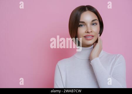 Strahlende junge Dame mit glänzenden Haaren in einem weißen Rollkragen, Hand auf Wange, vor einem leuchtend rosa Hintergrund Stockfoto