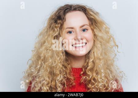 Helläugige junge Frau mit voluminösen lockigen blonden Haaren, die mit einem fröhlichen Lächeln in rotem Strick strahlt. Stockfoto