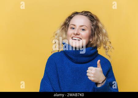 Überschwängliche junge Frau mit blonden lockigen Haaren, die ihre Daumen in einem gemütlichen blauen Rollkragenkragen gegen eine gelbe Wand hält. Stockfoto