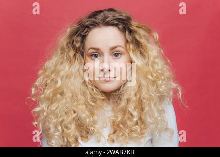 Blond mit lockigen Haaren und verspielter Neugier, in einem weißen Pullover auf rotem Hintergrund Stockfoto