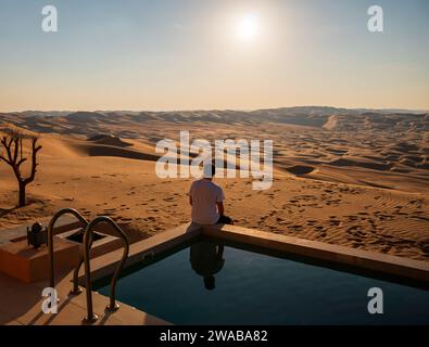 Ein Mann, der am Rand eines privaten Pools vor dem Hintergrund von Sanddünen und Wüstenlandschaft in der Wüste RUB'al Khali, Liwa, sitzt. Heller Sonnenschein Stockfoto