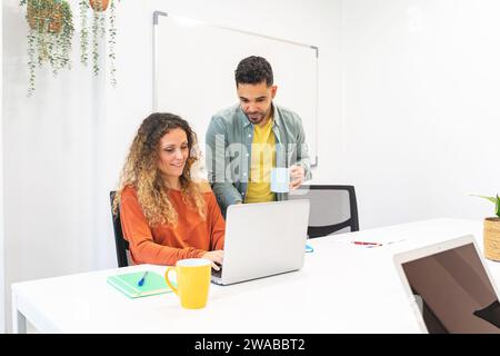 Zwei engagierte Kollegen arbeiten mit einem Laptop in einem lebhaften Büro zusammen, mit einem Whiteboard und Pflanzen im Hintergrund Stockfoto